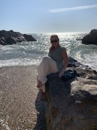 Full length of man sitting on rock at beach against sky