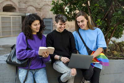 Young diverse people with lgbt rainbow flag using laptop and book outdoors.