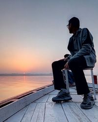 Man sitting on seat in sea against sky during sunset