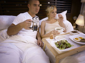 Couple with food and drink on bed in hotel room