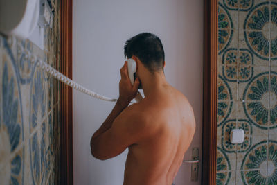 Rear view of shirtless man talking on phone while standing against wall at home