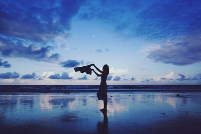 Silhouette woman standing while holding textile at beach against sky during sunset