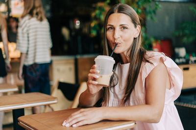A woman sitting in a cafe drinks iced coffee and looks at the camera.