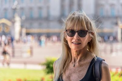 Mature blonde woman with glasses looking at the camera. behind out of focus buckingham palace.