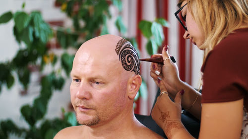 Tattoo master, mehendi artist makes drawing of henna tattoo on the scalp of bald caucasian man 