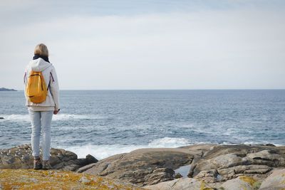 Rear view of woman with backpack standing on rocky beach against sky