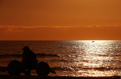 Silhouette man sitting on beach against orange sky