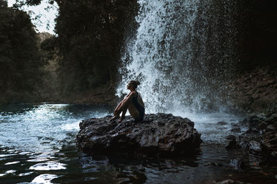 Side view of young woman sitting by waterfall on rock