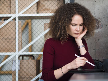 Beautiful woman with curly hair with pen in hand working with tablet while sitting at table in cafe.