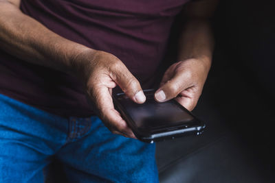 Senior ethnic male in glasses relaxing on couch in living room and reading messages on smartphone