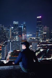 Rear view of man sitting on building terrace against illuminated cityscape at night