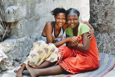 Portrait of female friends smiling while sitting against wall