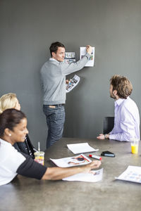 Mid adult businessman sticking photograph on wall while colleagues looking at him in office