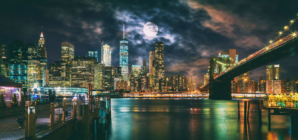 Illuminated modern buildings by river against sky in city at night