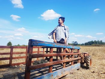 Man standing on abandoned cart at field against blue sky