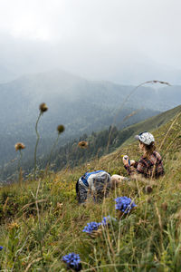 Young woman sitting on meadow in mountain valley next to backpack drinking water  on hike landscape 