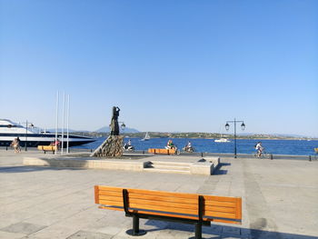 Empty bench on walkway by sea against clear blue sky