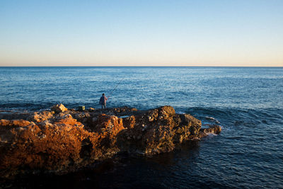 Rear view of man fishing on rocky shore against clear sky