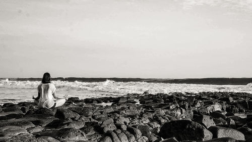 Rear view of woman sitting on rock by sea against sky