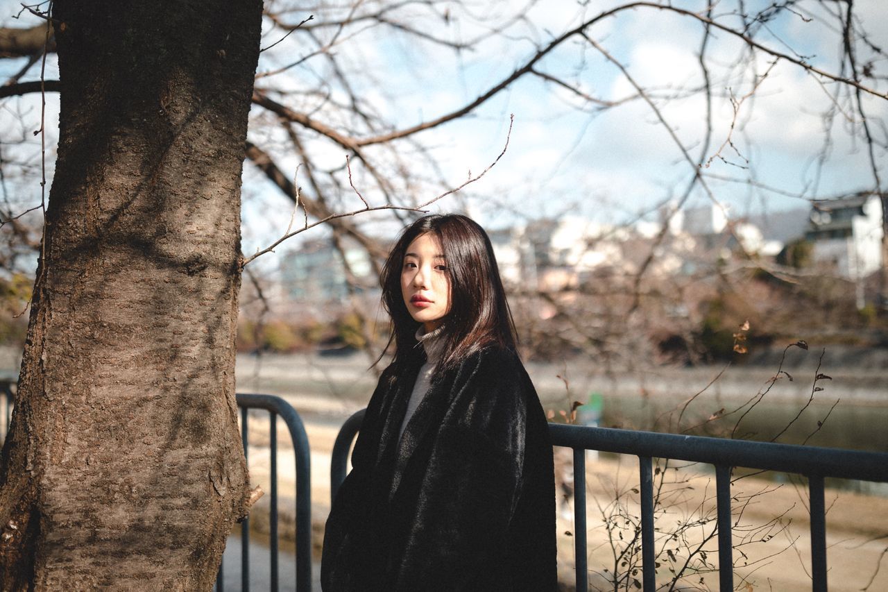 tree, one person, young adult, standing, young women, leisure activity, real people, lifestyles, portrait, hairstyle, plant, looking at camera, hair, long hair, railing, women, focus on foreground, tree trunk, trunk, outdoors, warm clothing, beautiful woman, contemplation