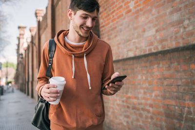 Smiling young man holding coffee while using smart phone against wall