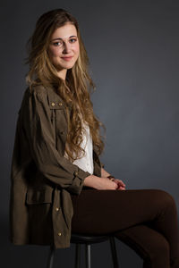 Portrait of young woman with blond hair sitting on stool against black background