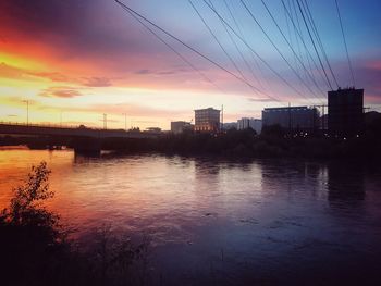 Silhouette buildings by river against sky at sunset