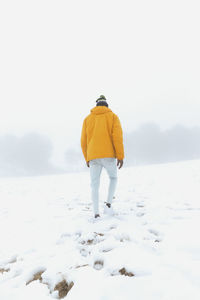 Back view of anonymous ethnic male in outerwear and jeans walking on snow and leaving footprints in winter meadow