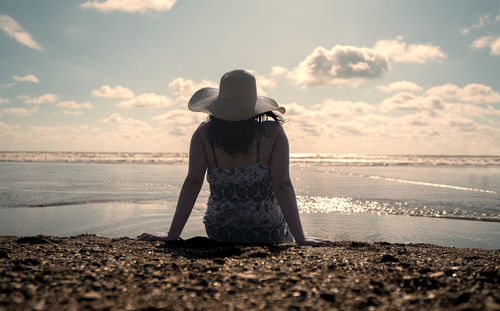 Woman sitting alone at the edge of the beach seen from behind, wearing a hat  during sunset