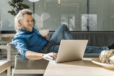 Mature man with a beard smiling and working with his laptop on a sofa