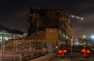 Illuminated construction site by buildings against sky at night