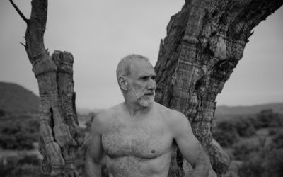 Portrait of shirtless adult man with tree trunk against sky