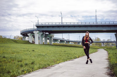 Full length portrait of fashionable young woman standing on footpath amidst field with bridge in background