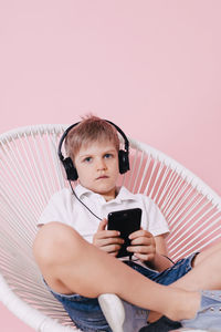 Portrait of boy listening music while using phone on chair against colored background