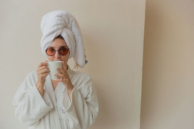 A womaa woman with glasses in a bathrobe and a towel at the hotel drinks coffee from a white cup.