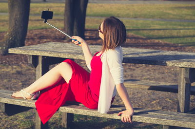 Side view of sensuous young woman holding monopod while taking selfie through smart phone on picnic table in park