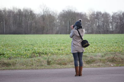 Rear view of woman photographing while standing on road by field