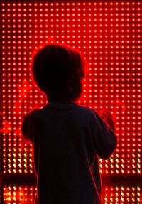 Rear view of boy standing in front of illuminated glass