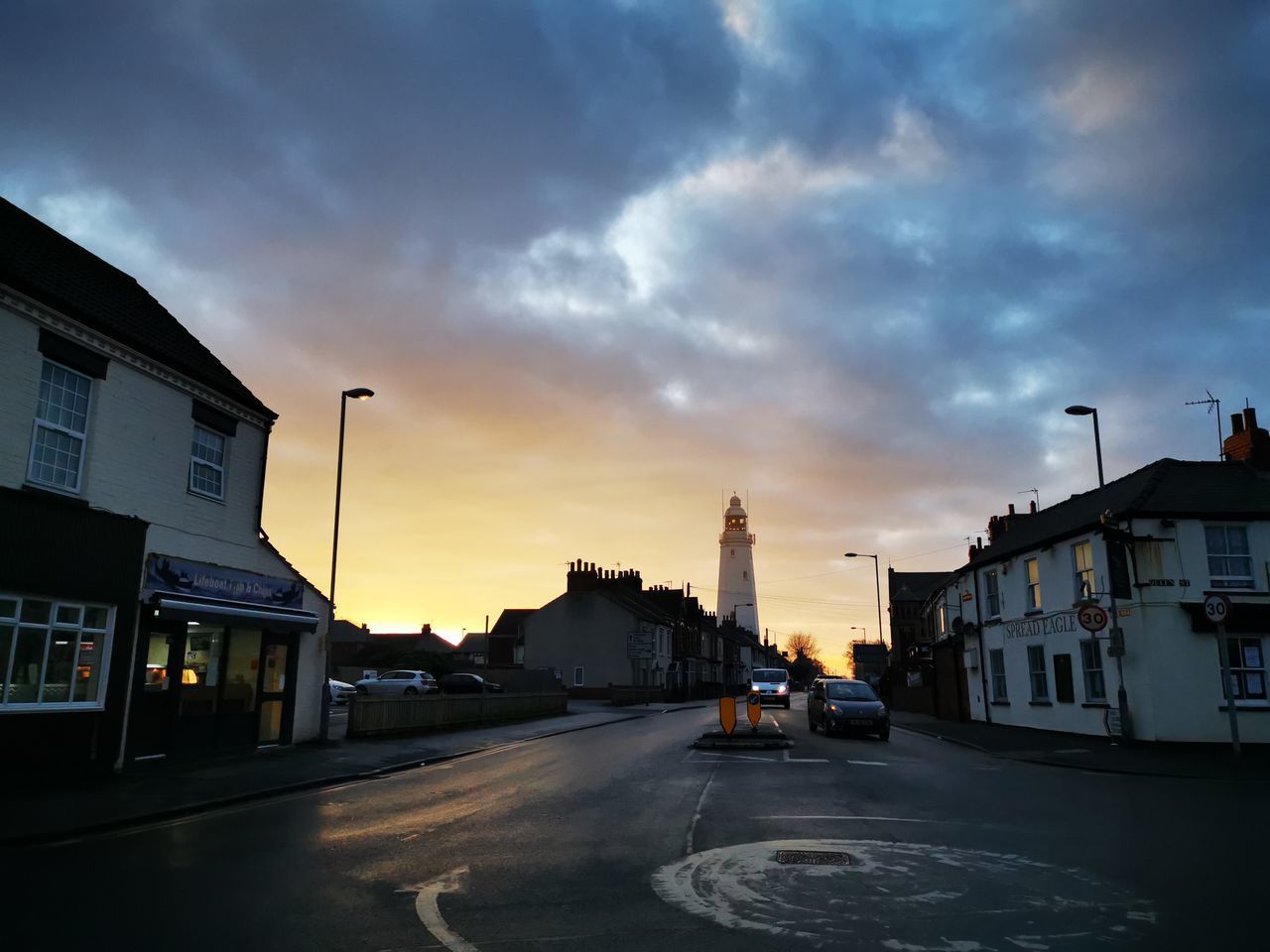 CARS ON ROAD BY BUILDINGS AGAINST SKY DURING SUNSET