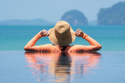 Woman wearing swimsuit standing in infinity swimming pool looking at ocean on summer vacation