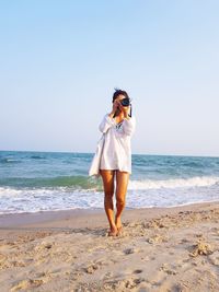 Full length of young woman photographing at beach against clear sky