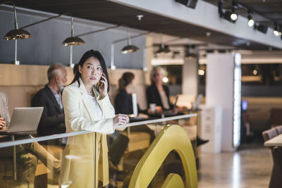 Female entrepreneur talking on mobile phone while standing by railing with colleagues in background at office