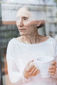 Thoughtful mature woman looking through window while holding coffee mug at home