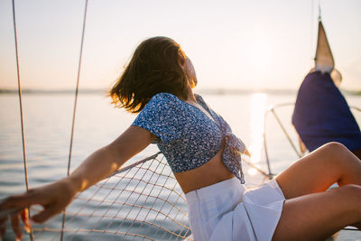 Side view of unrecognizable young stylish female traveler with dark hair in trendy crop top and skirt admiring sea while sitting on sailboat during cruise under sunset sky