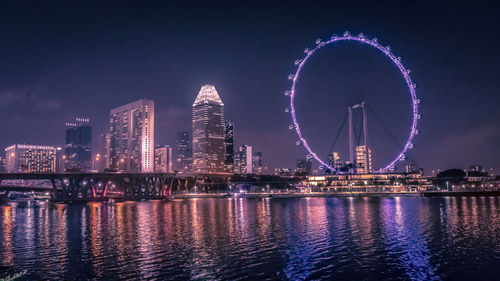 Illuminated ferris wheel by river against sky at night