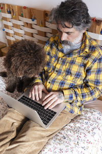 Man sitting on bed with dog and working on laptop, enjoying remote work and pet companionship
