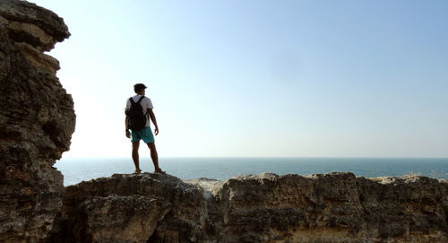 Rear view of man standing on cliff by sea against clear sky