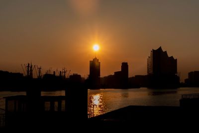 Silhouette buildings by river against sky during sunset