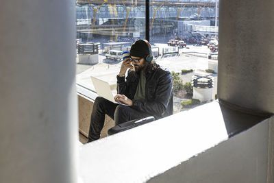 Young hipster guy in black jacket and hat with headphones drinking takeaway coffee and using laptop while sitting against glass wall in airport lounge