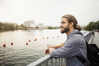 Thoughtful smiling man standing at railing by river in city against clear sky